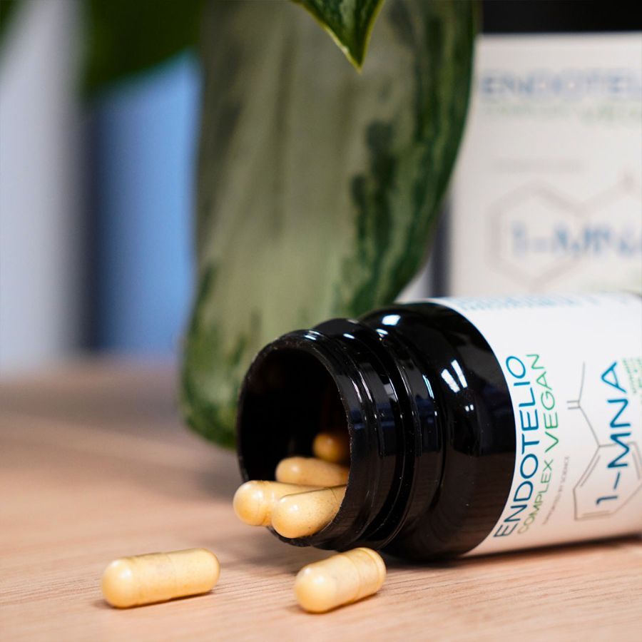 Flacone marrone con capsule cadute su una superficie di legno. Scritta: Endotelio Complex Vegan, 1-MNA. Alcune capsule accanto.