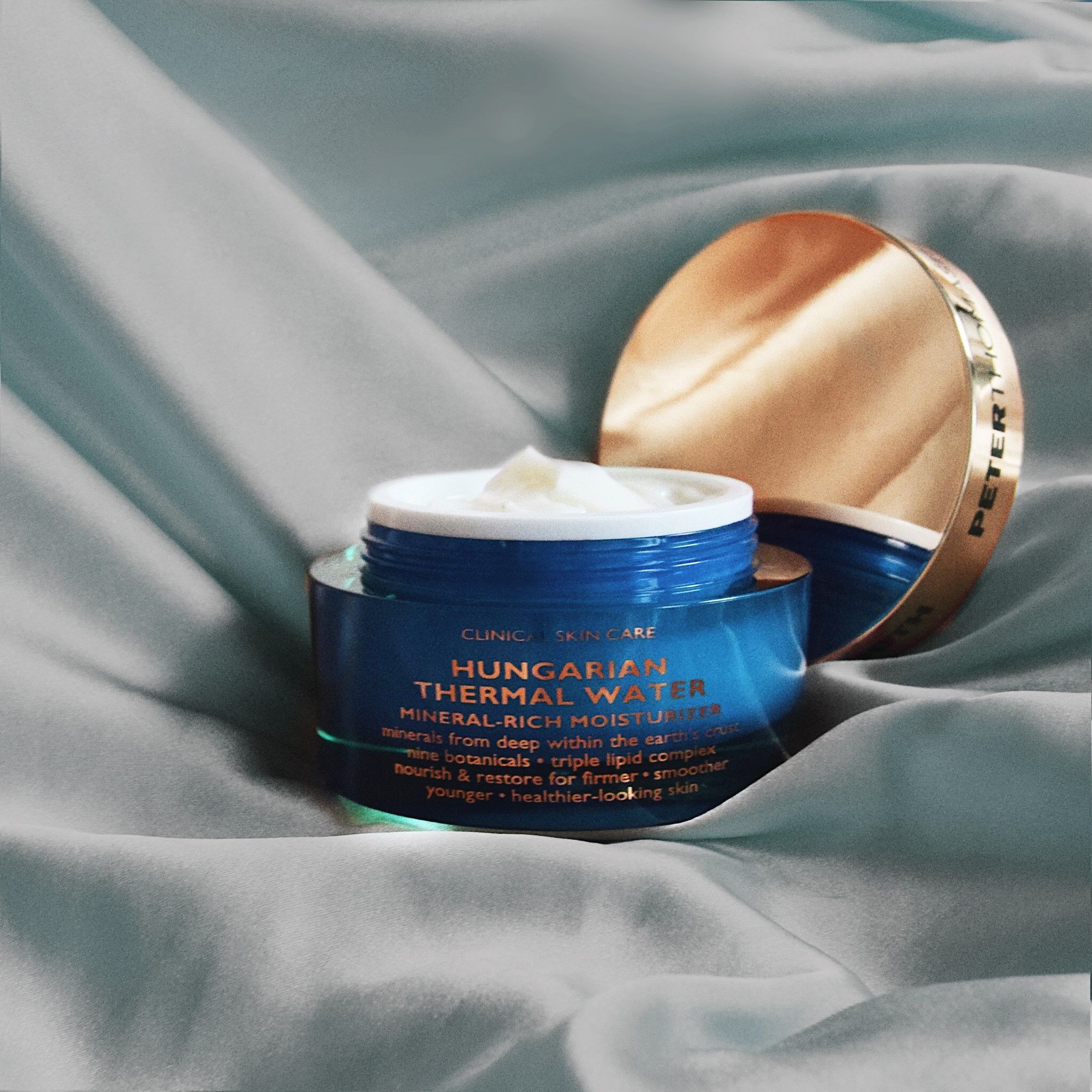 Vaso aperto con coperchio dorato su tessuto azzurro. Nome del prodotto e testo visibili. Crema all'interno.