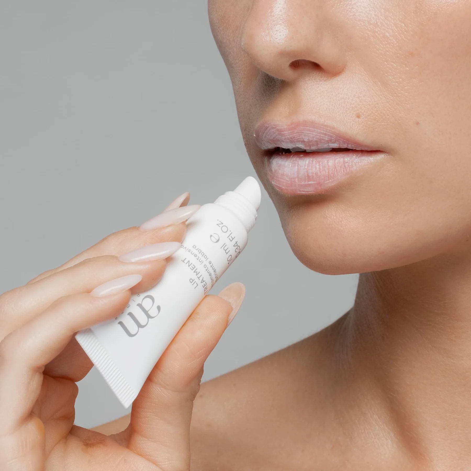Una persona tiene in mano un tubo bianco per il trattamento labbra. Il tubo viene applicato sulle labbra. Il tubo riporta "am" e "LIP TREATMENT".