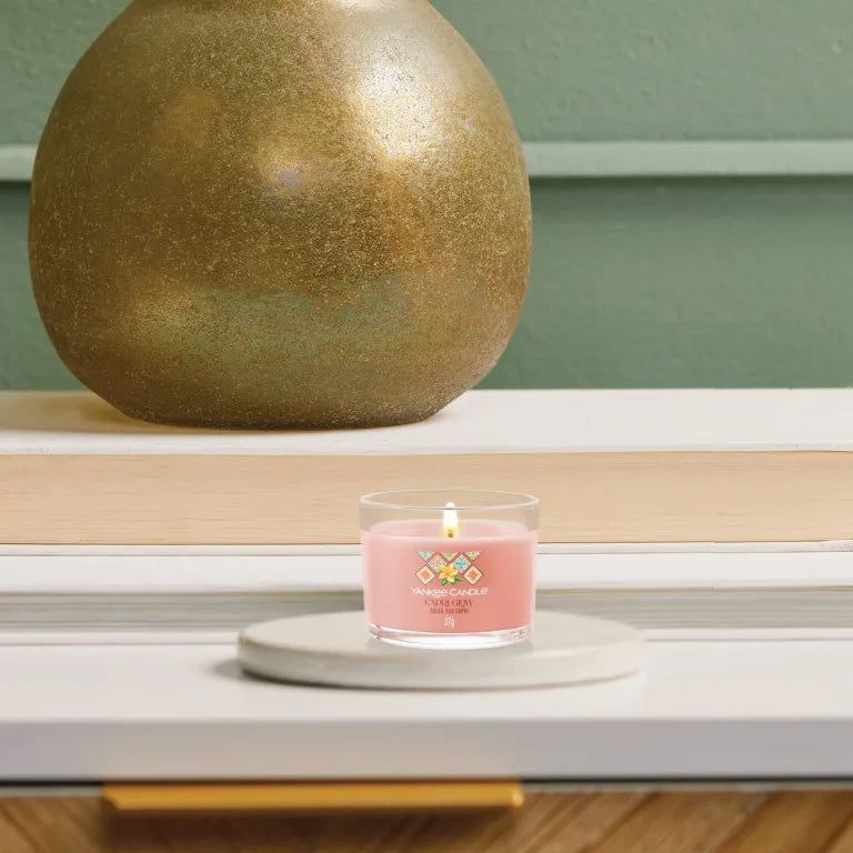 Candela rosa in contenitore di vetro su sottobicchiere. Scritta: Capri Glow, Soleil sur Capri. Candela accesa.