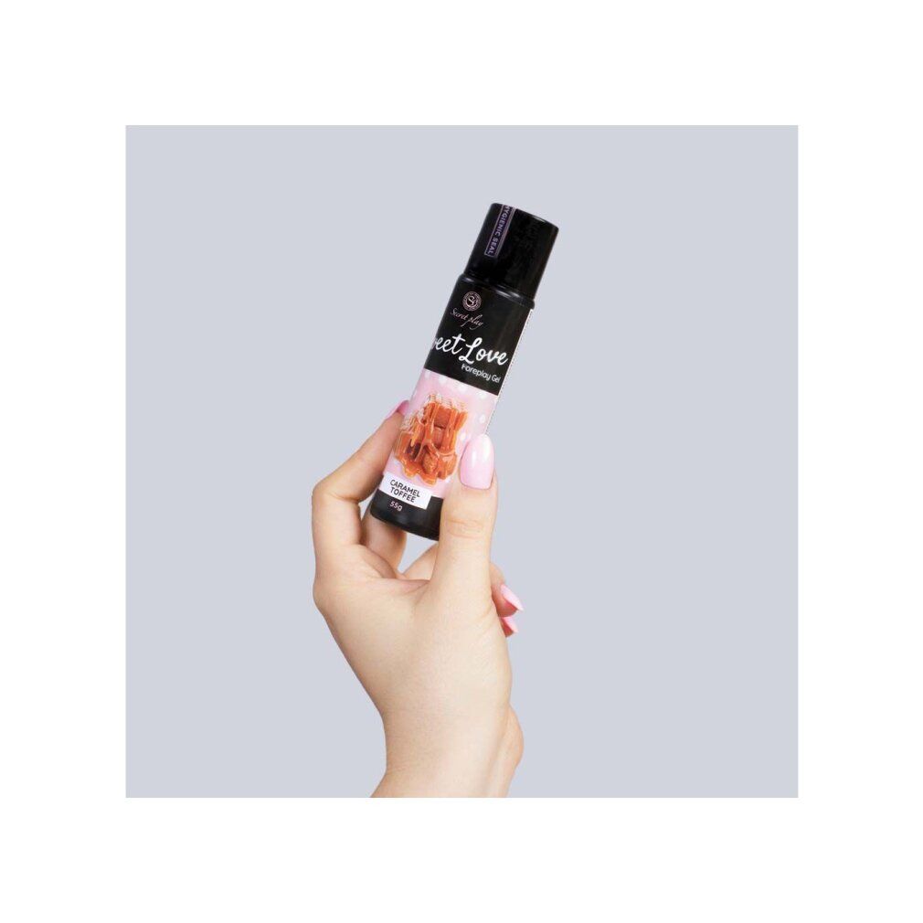Mano che tiene un flacone nero. Scritta: Sweet Love Foreplay Gel. Illustrazione di caramello mou. 55g. Sfondo rosa.