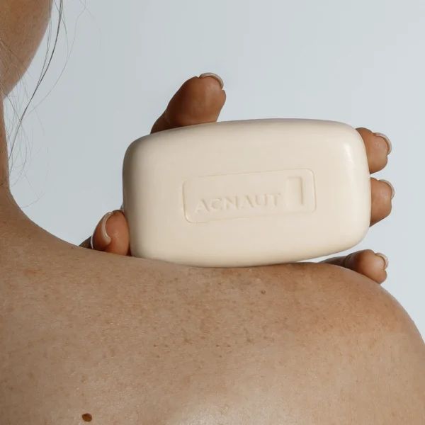 Una persona tiene in mano un pezzo di sapone rettangolare. Sul sapone c'è scritto "Acnaut". La pelle è visibile.
