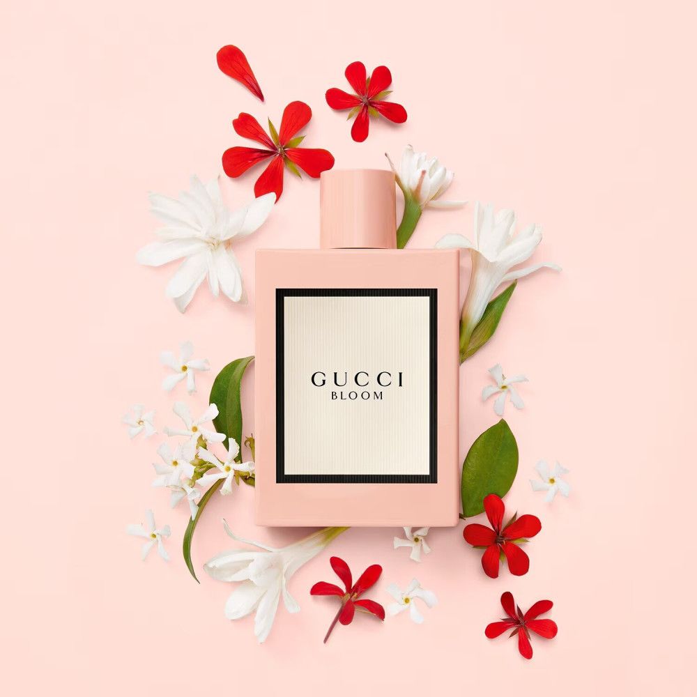 Flacone rosa circondato da fiori bianchi e rossi su sfondo rosa. Sul flacone: Gucci Bloom, etichetta bianca con cornice nera.