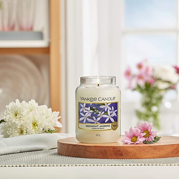 Candela profumata in vetro su vassoio di legno. Adesivo con motivo floreale e scritta Midnight Jasmine. Composizioni floreali sullo sfondo.