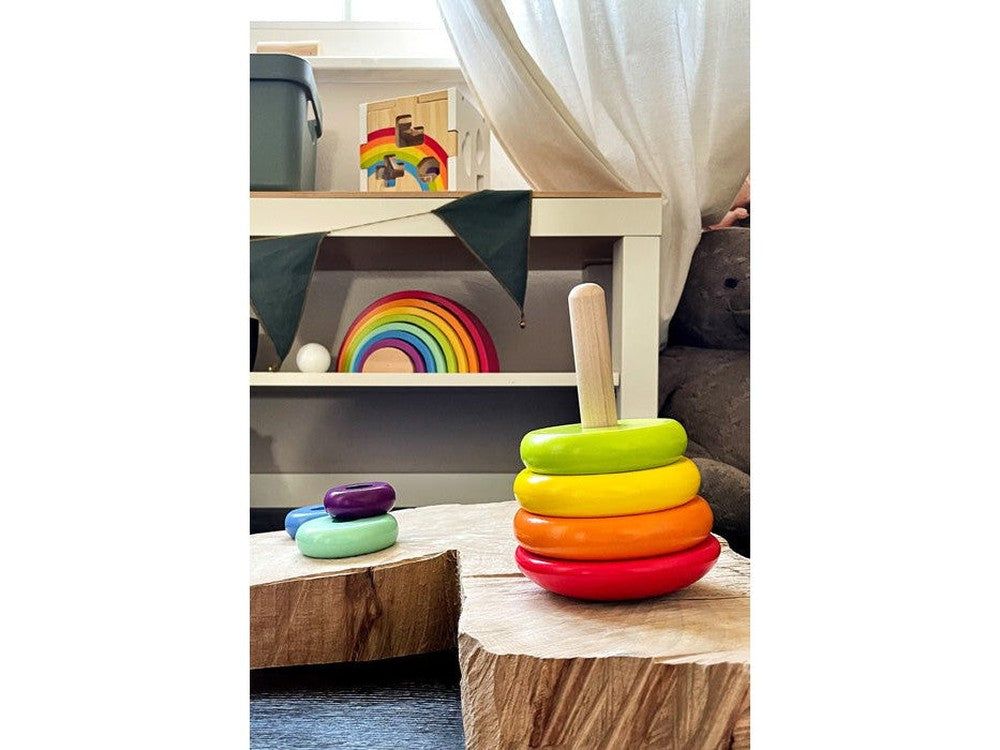 Torre impilabile con asta in legno e 6 anelli su blocchi di legno. Colori: rosso, arancione, giallo, verde, turchese, viola. Arcobaleno sullo sfondo.