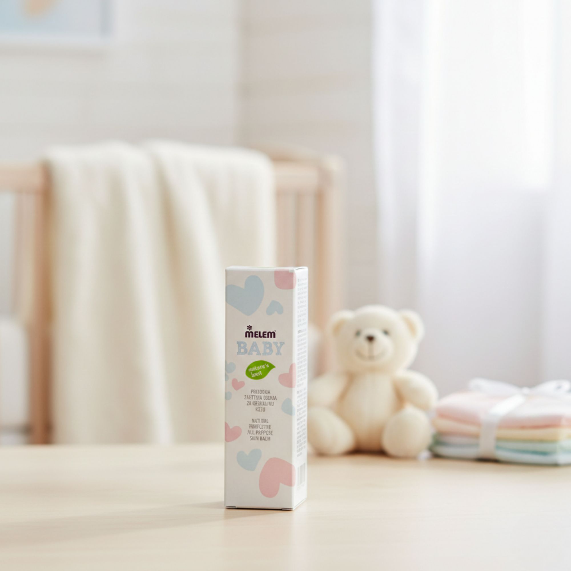 Confezione "Melem Baby" su tavolo. Testo: "Natural Protective All-Purpose Skin Balm". Sfondo: lettino, orsacchiotto.