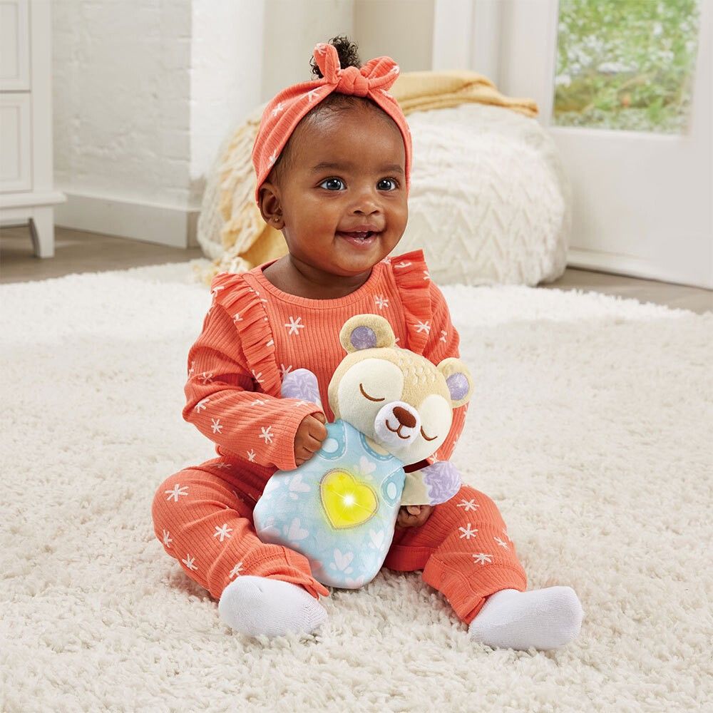 Bambino tiene orsetto con cuore. Orsetto beige, vestito blu con cuore. Cuore luminoso. VTech.