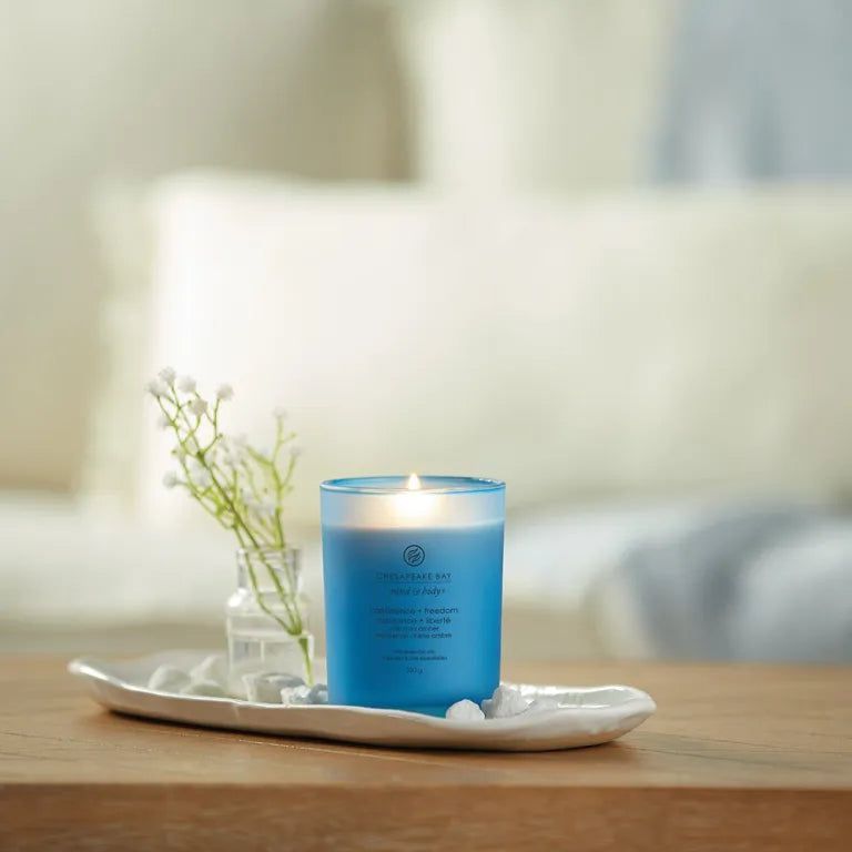 Candela blu accesa su vassoio. Accanto: vaso con fiori e pietre. Sfondo: divano.