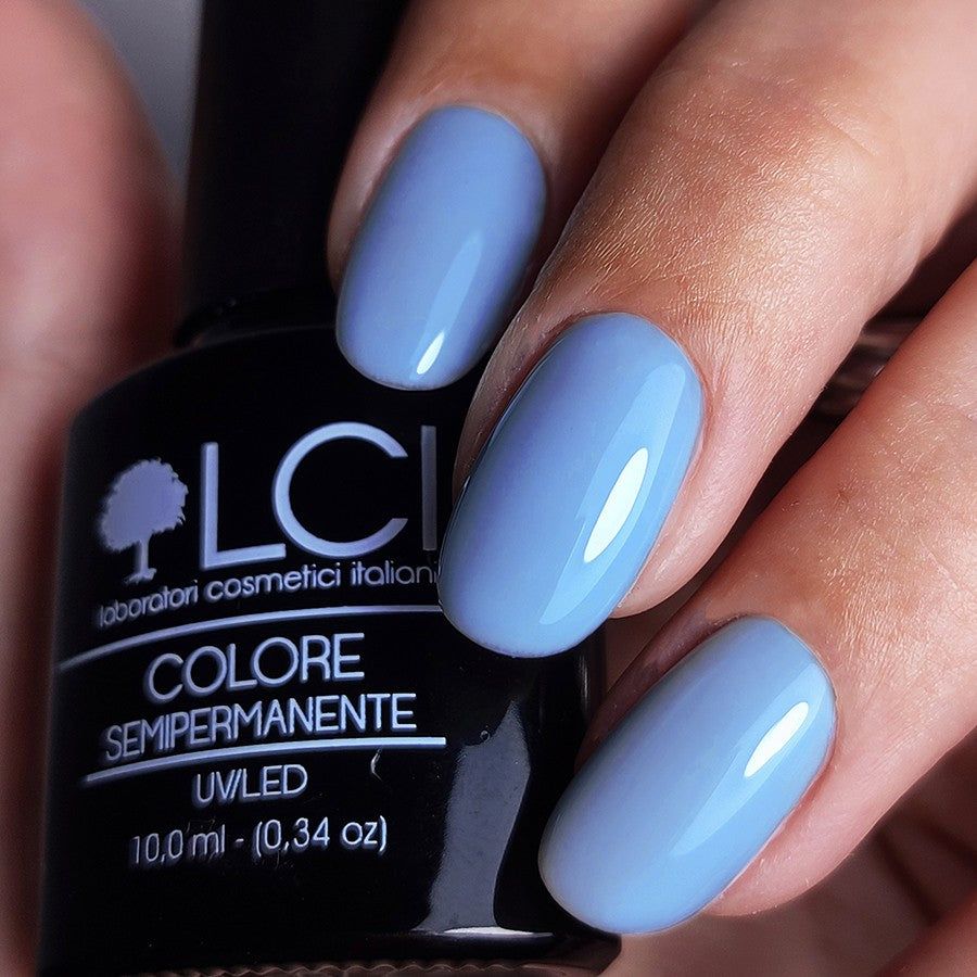 Mano con unghie smaltate di azzurro chiaro. Flacone di smalto nero con testo bianco. "COLORE SEMIPERMANENTE" visibile.