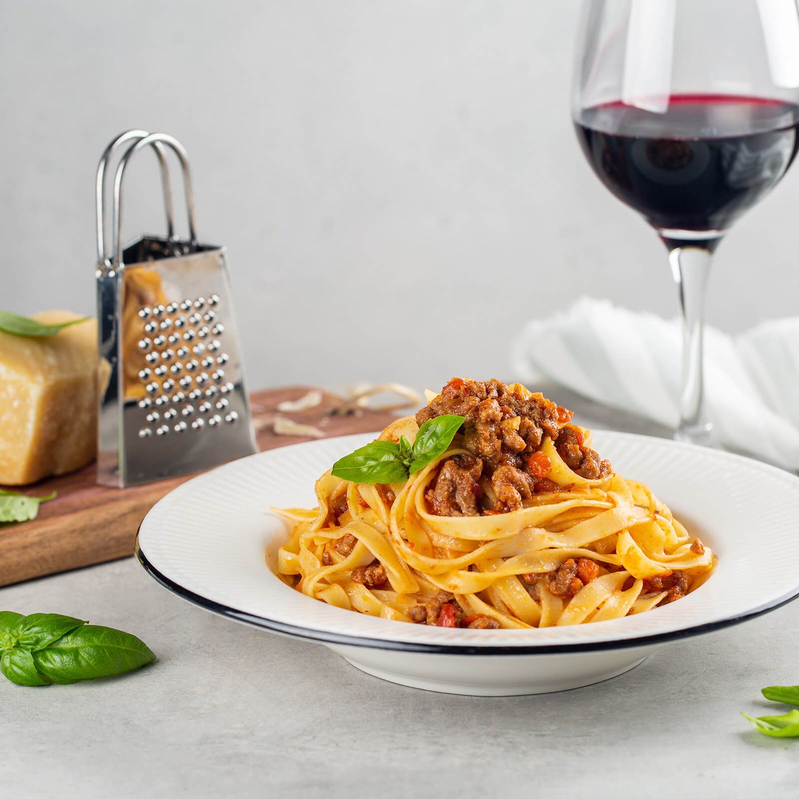 Tagliatelle con ragù, foglia di basilico. Formaggio, grattugia, vino e tagliere sullo sfondo.