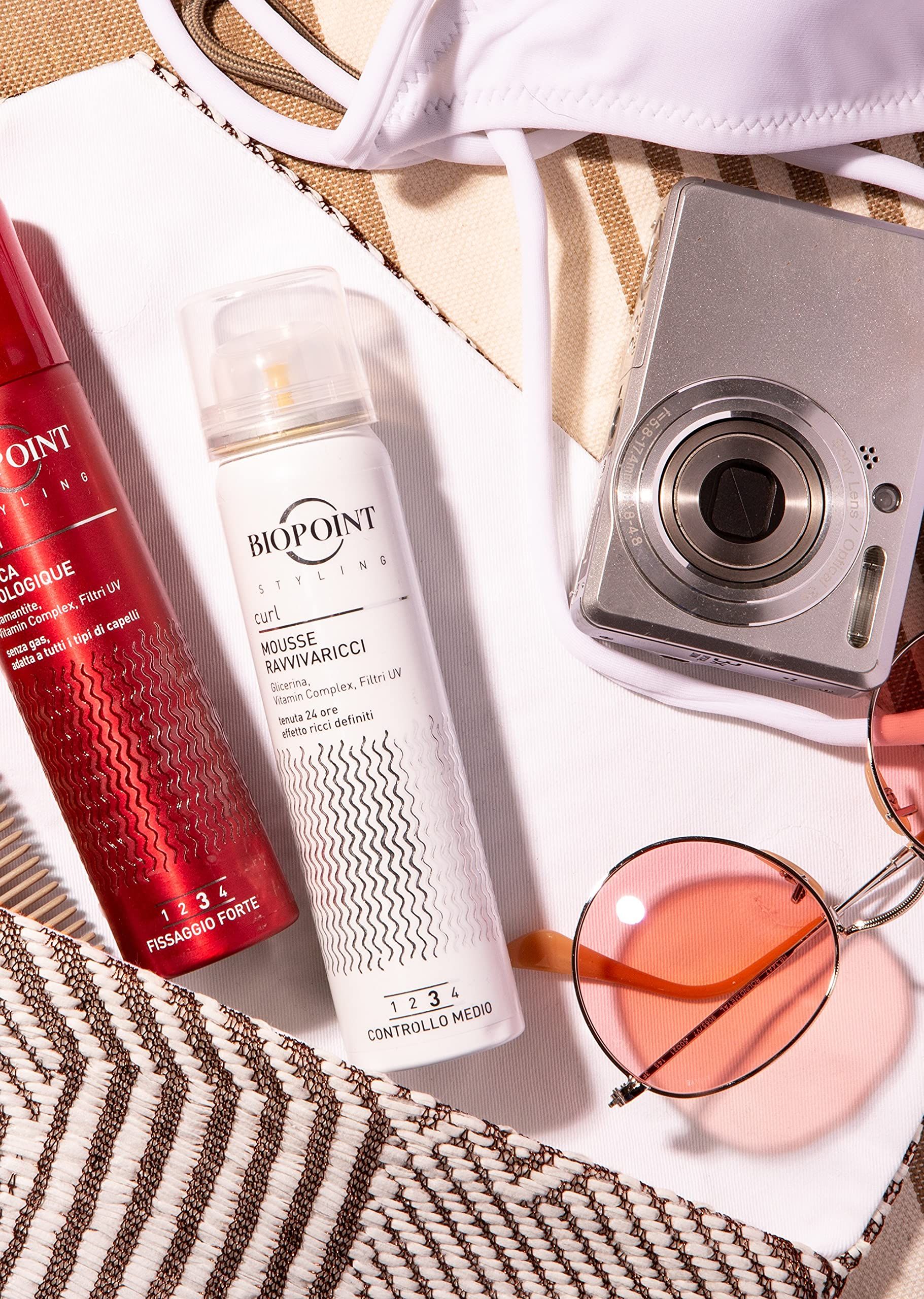 Due spray, tra cui Mousse Biopoint, accanto a fotocamera, occhiali da sole e bikini. Spray rossi e bianchi con testo.