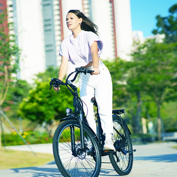 E-bike con persona. La persona indossa abiti bianchi. Guida su strada. Edifici e alberi sullo sfondo.