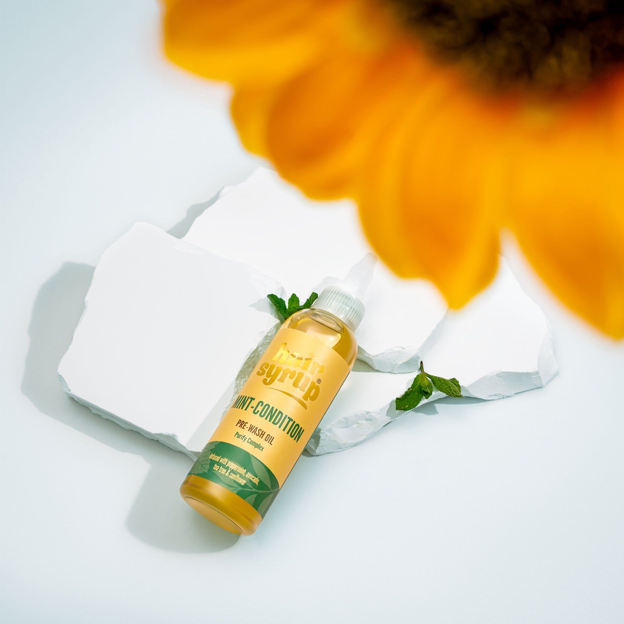 Flacone giallo "Hair Syrup Mint Condition" su pietre bianche, menta e fiore sullo sfondo.