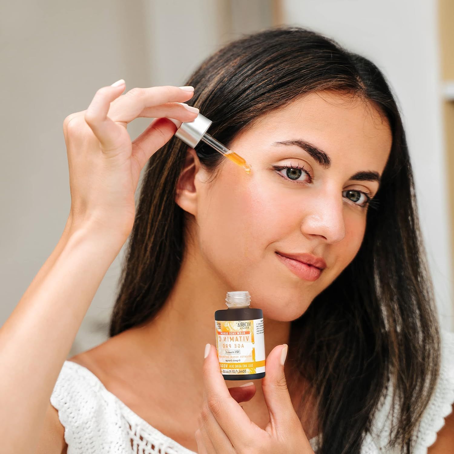 Donna che tiene un flacone di siero. Gocce sulla pelle. Scritta: Vitamin C Age Pro.