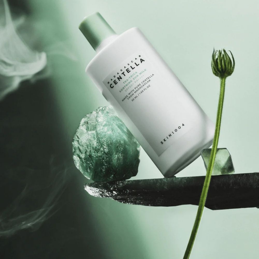 Flacone Madagascar Centella Tea-Trica Soothing Sun Milk SPF50+ PA++++ su pietra con cristalli verdi e fumo.