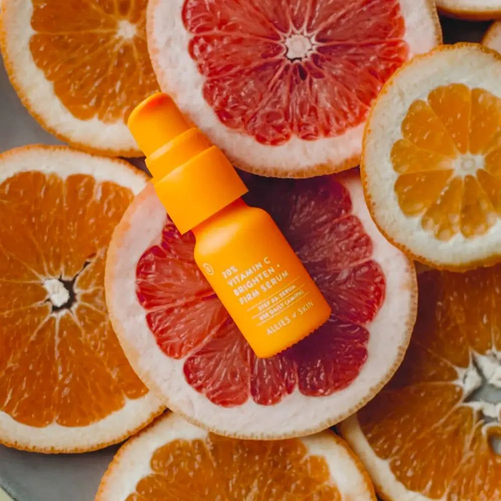 Flacone di siero arancione su fette di pompelmo e arancia. Scritta: 20% Vitamin C Brighten + Firm Serum. Marchio: Allies of Skin.