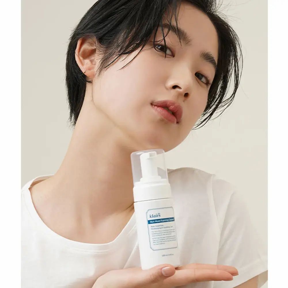 Persona che tiene in mano un flacone bianco Klairs Rich Moist Foaming Cleanser. Etichetta blu con testo. Tappo trasparente. Pelle chiara.