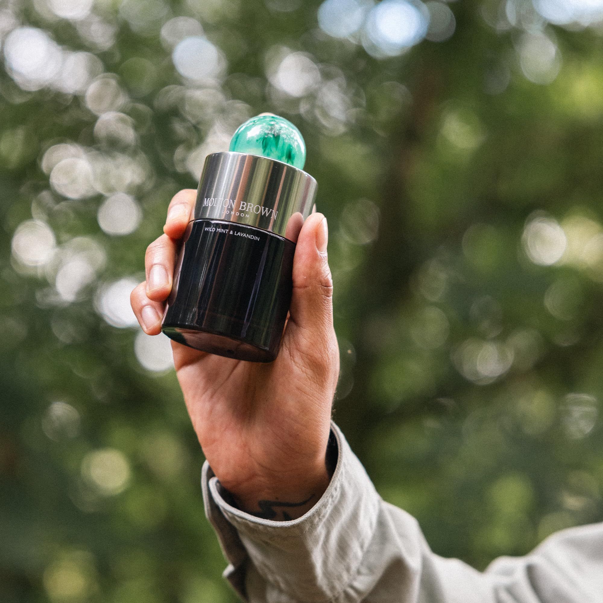 Mano che tiene un flacone di vetro scuro con tappo verde. Scritta: Wild Mint & Lavandin. Sfondo: foresta sfocata.