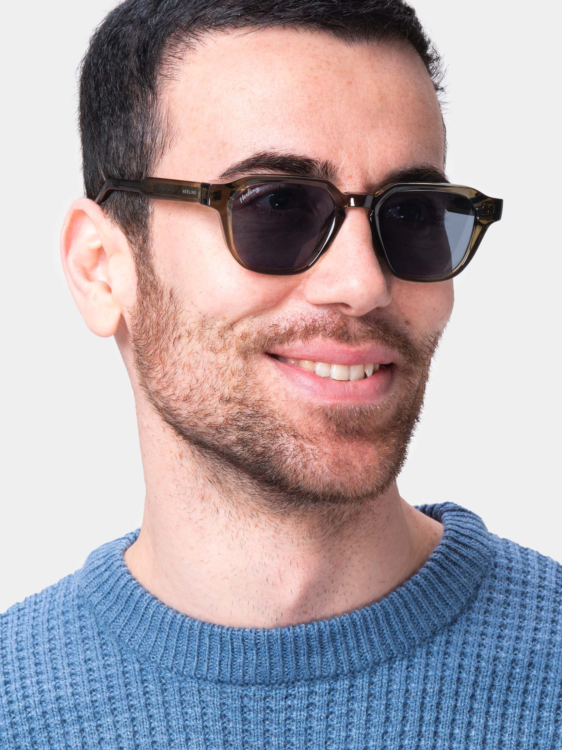 Uomo con occhiali da sole verdi scuro. Lenti quadrate. Logo Herling sulla stanghetta. Maglione blu.