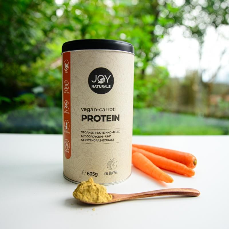 Barattolo accanto a carote e polvere su cucchiaio. Scritta: JOY NATURALS, vegan-carrot:PROTEIN, 605g. Complesso proteico vegano con estratto di Cordyceps.