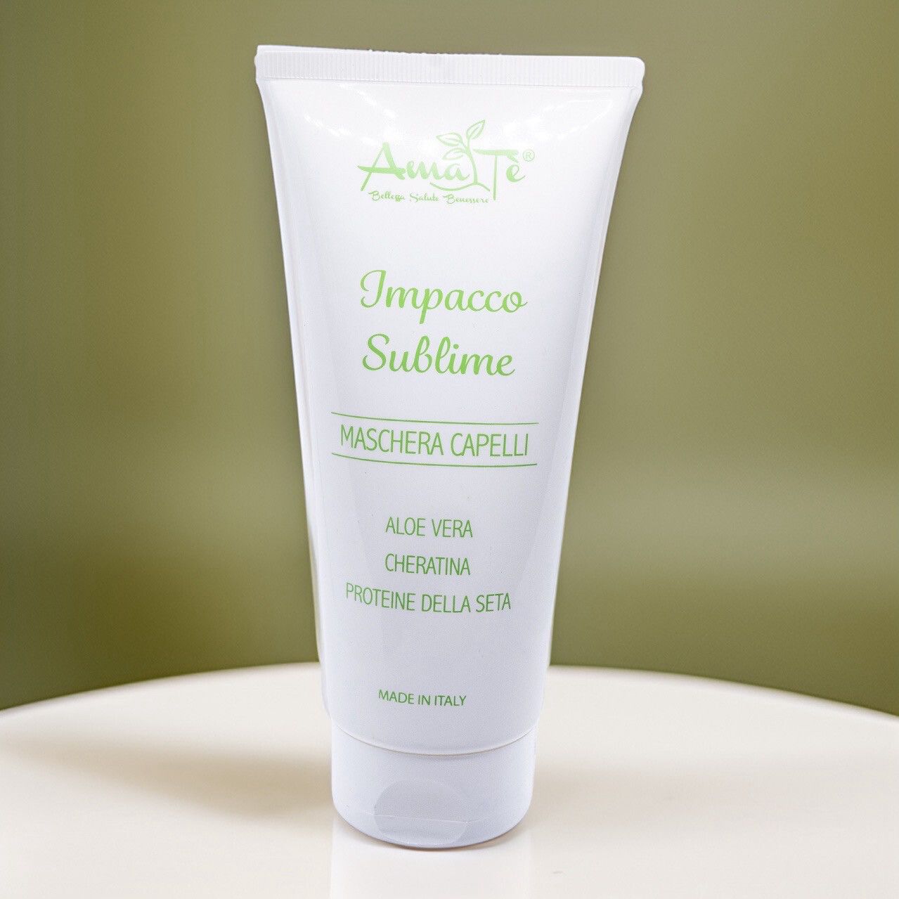 Tubo bianco con testo verde: Amatè, Impacco Sublime, Maschera Capelli. Aloe Vera, Cheratina, Proteine della seta. Su tavolo bianco, sfondo verde.