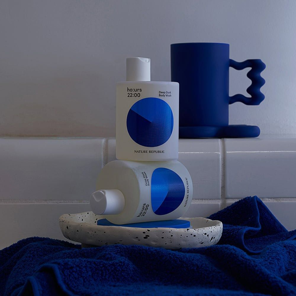 Due flaconi di bagnoschiuma, bianco con design blu. Marchio: NATURE REPUBLIC. Testo: ho:urs 22:00 Deep Dark Body Wash. Su un vassoio e un asciugamano.