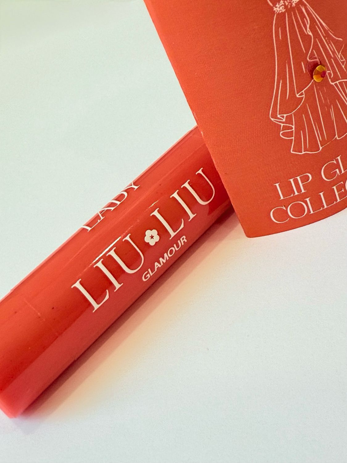 Rossetto rosso e confezione arancione. Scritta "LADY LIU LIU GLAMOUR" e disegno di un vestito.