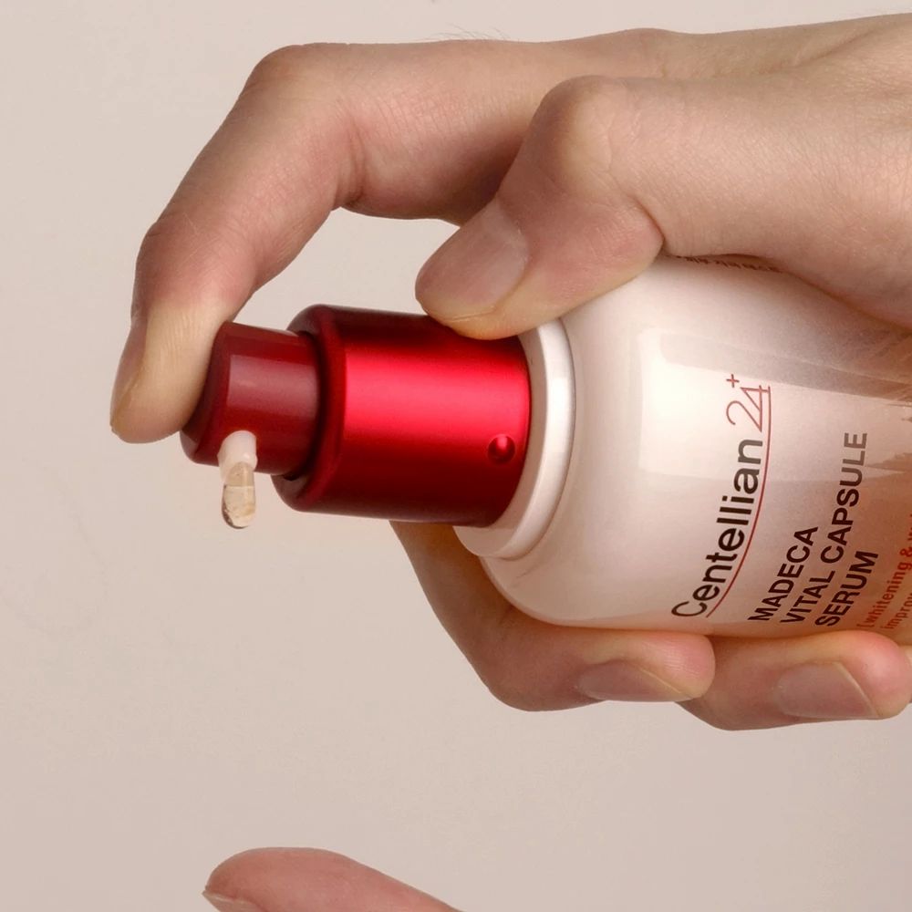 Mano che preme il siero dalla bottiglia. Testa della pompa rossa. Nome del prodotto e marchio visibili.