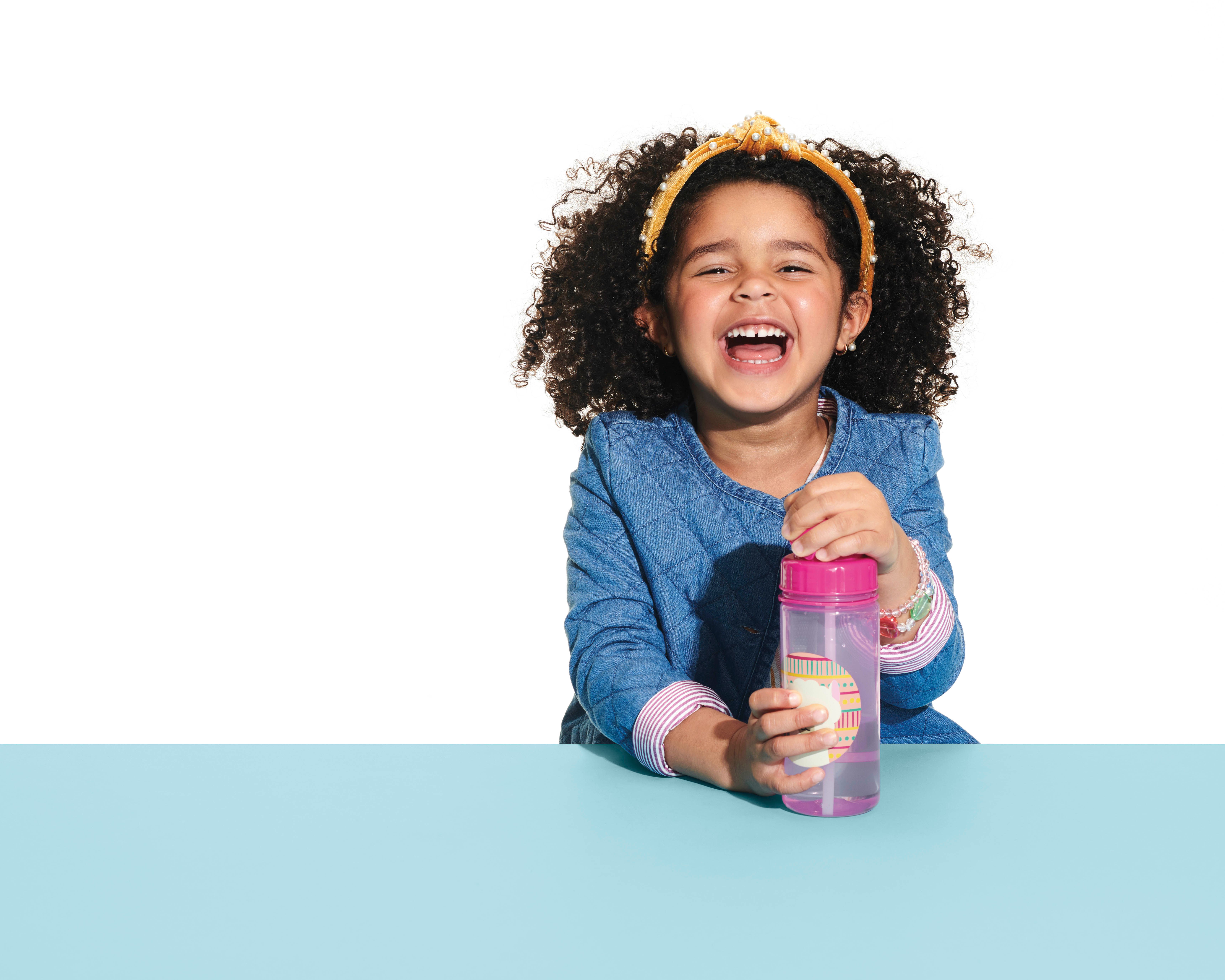 Bambina sorridente tiene in mano una borraccia rosa con motivo lama. La bambina indossa una camicia di jeans e una fascia. Piano del tavolo blu.
