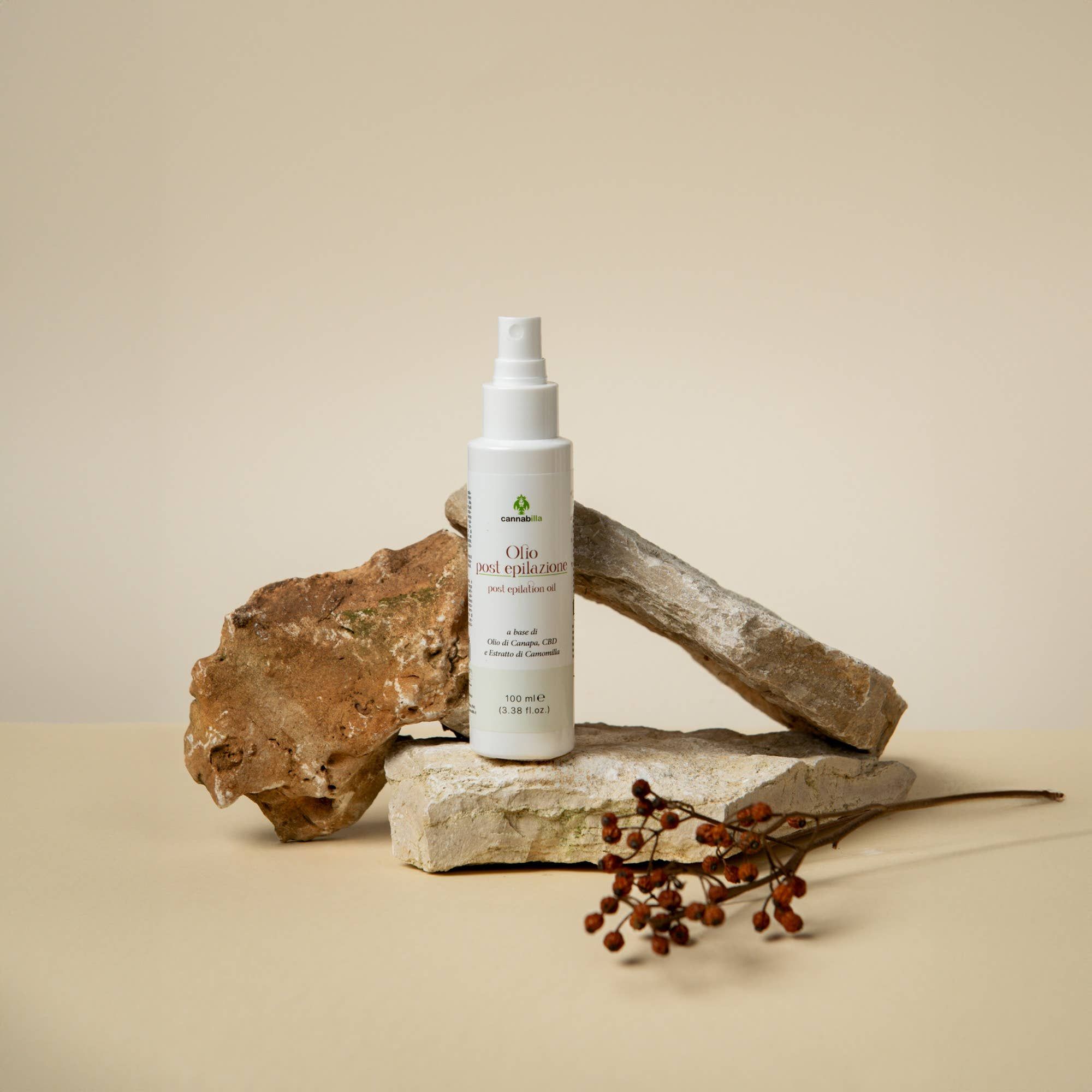Flacone spray bianco su pietre. Scritta: Olio post epilazione. Accanto: ramo con bacche rosse. Sfondo: beige.