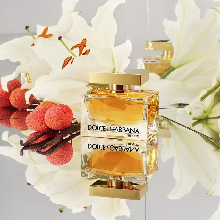 Flacone in vetro con tappo dorato, circondato da gigli e litchi. Scritta: Dolce & Gabbana, the one. Riflesso.