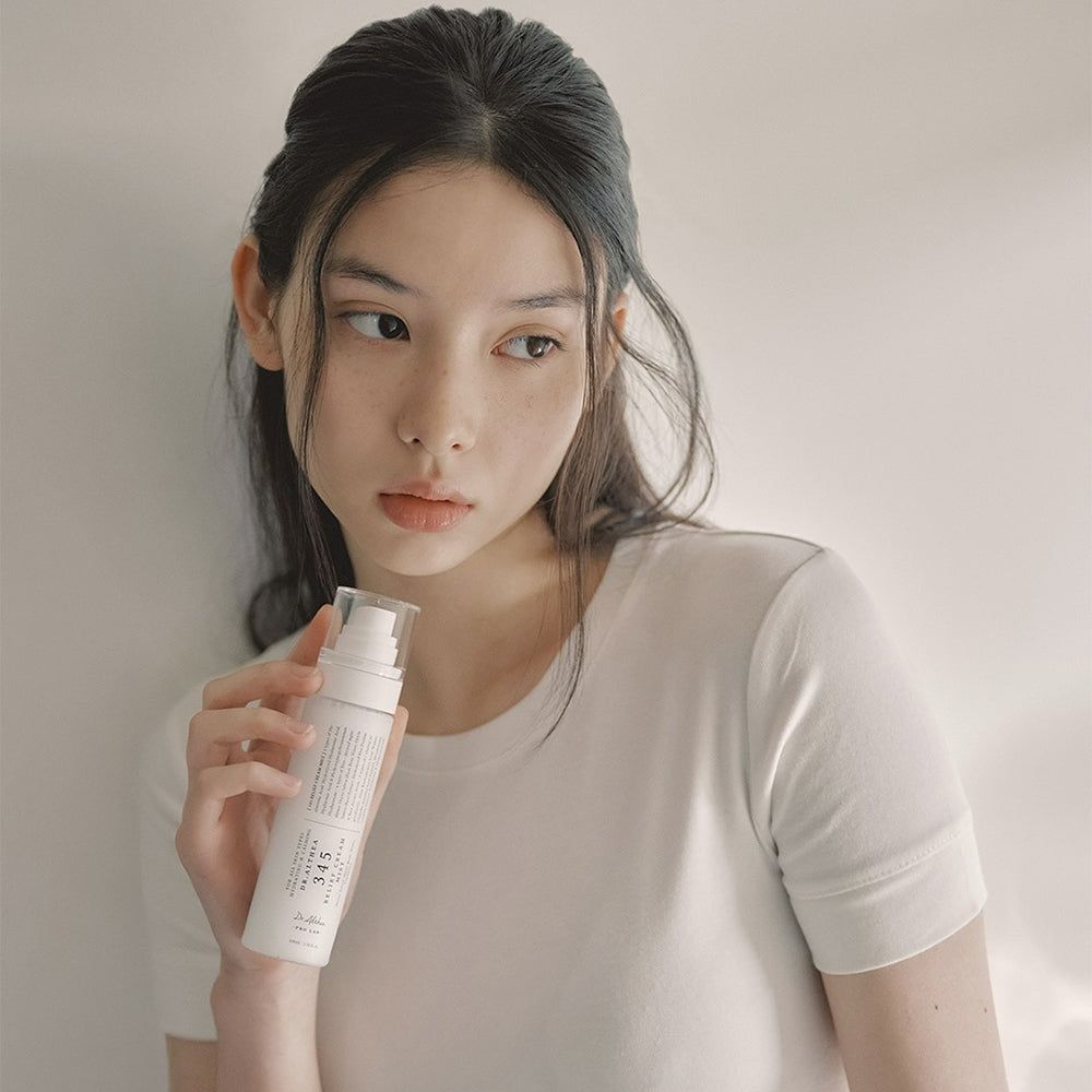 Una donna con i capelli scuri tiene una bottiglia spray bianca di Dr. Althea 345 Relief Cream Mist.