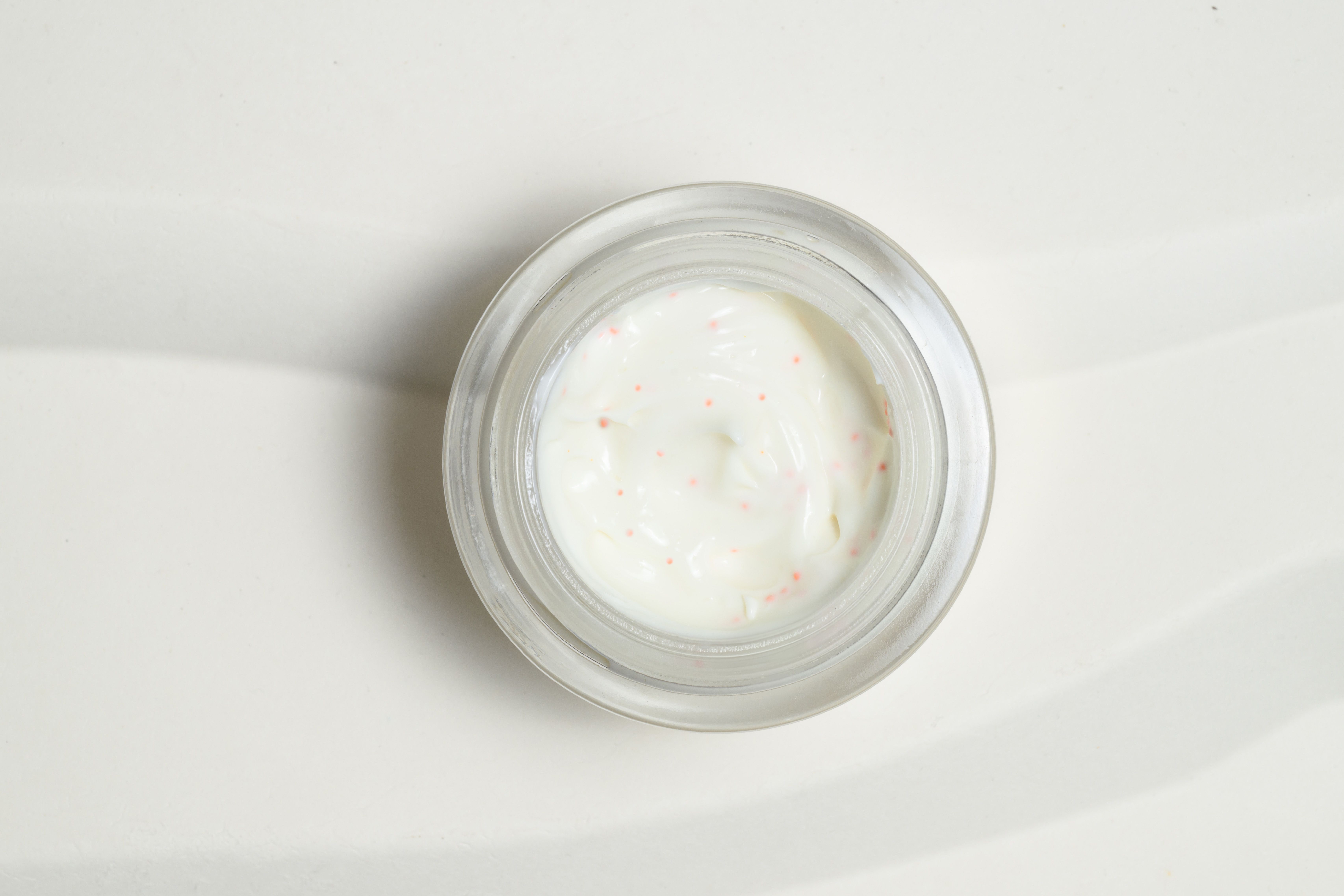 Crema in un contenitore di vetro. La crema è bianca con piccole particelle rosse. Il contenitore è in vetro.