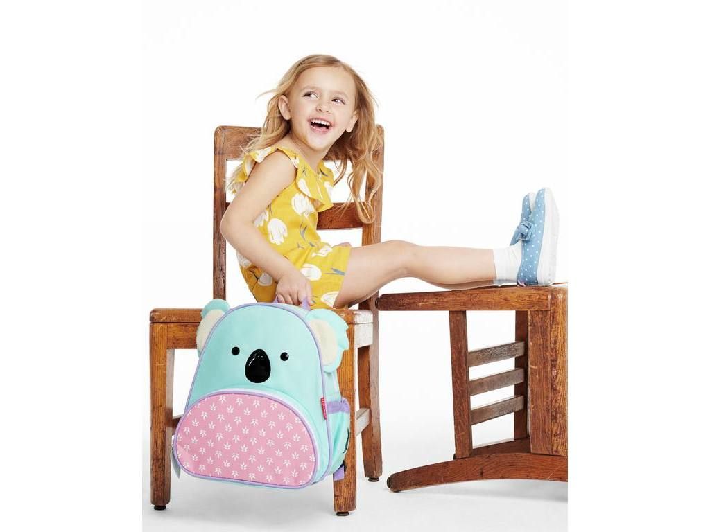 Bambina seduta su una sedia con zaino koala. Lo zaino ha orecchie, occhi e fondo rosa. Marchio Skip Hop.