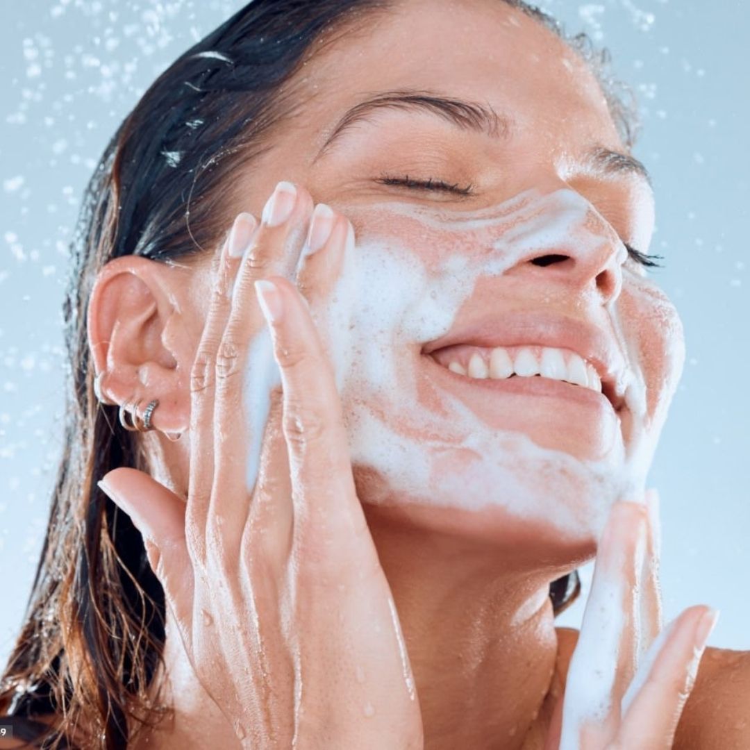 Donna che si lava il viso con schiuma bianca. L'acqua schizza. Sorride.