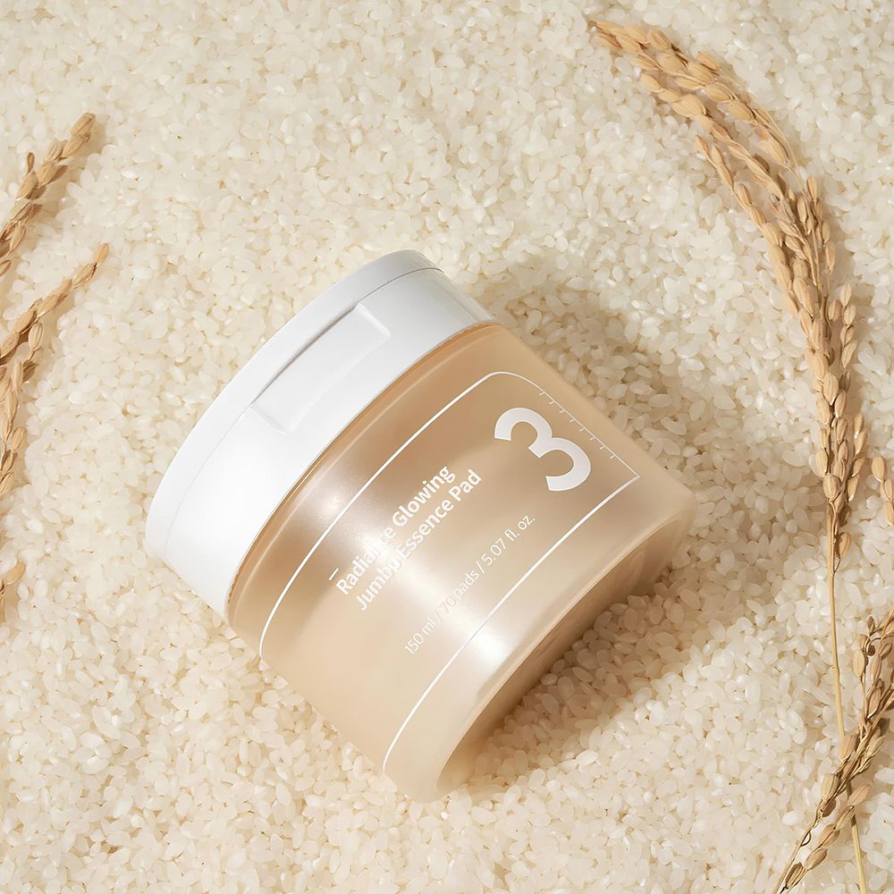 Barattolo beige con coperchio bianco. Scritta: 3, Radiance Glowing Jumbo Essence Pad. Posizionato su riso e spighe di grano.
