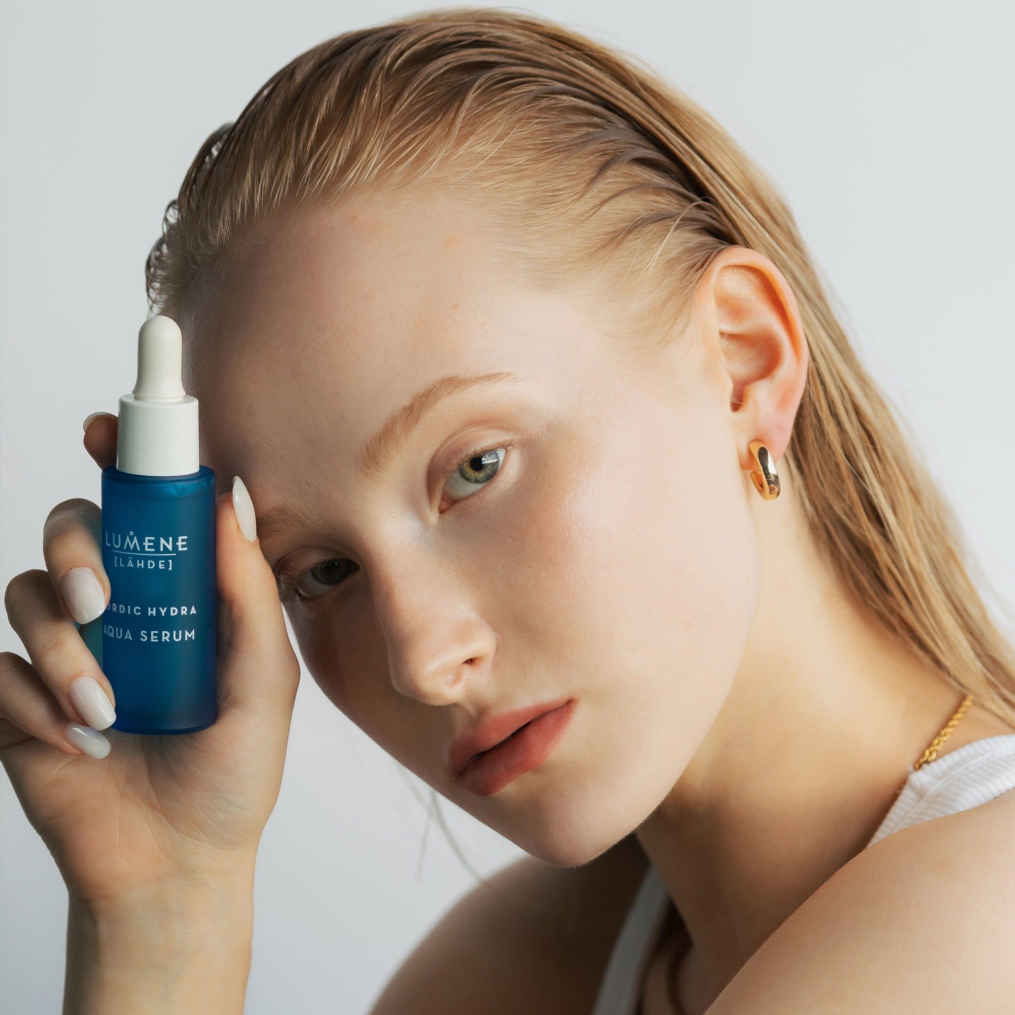 Donna con flacone di siero. Scritta: Lumene [LAHDE] Nordic Hydra Aqua Serum. La donna indossa orecchini d'oro.