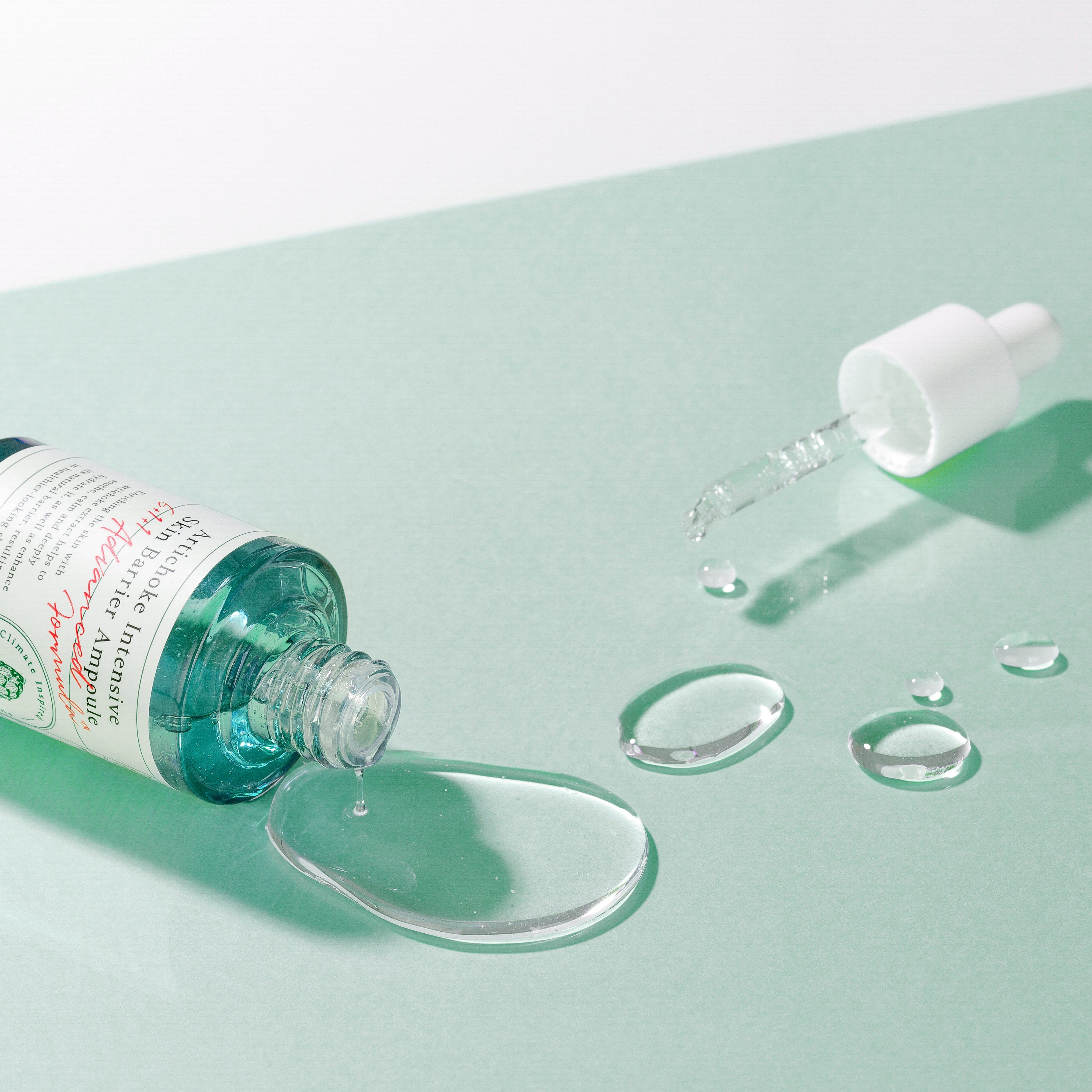 Flacone di siero e siero su una superficie. Scritta: Artichoke Intensive Skin Barrier Ampoule. AXIS-Y. Siero versato.
