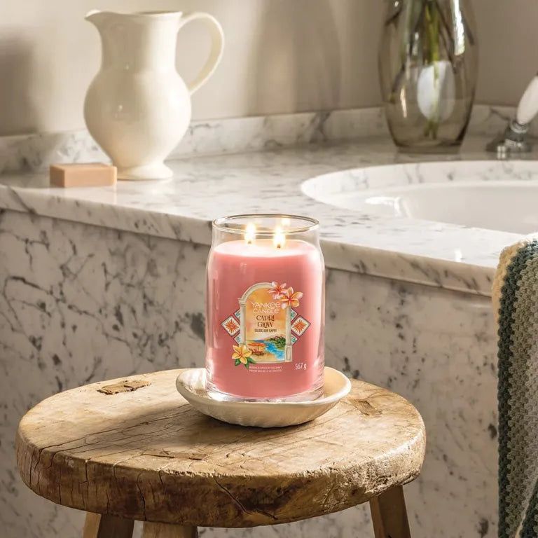 Candela rosa in vetro, due stoppini, su sottobicchiere. Scritta: Capri Glow. Su sgabello. Sfondo bagno.