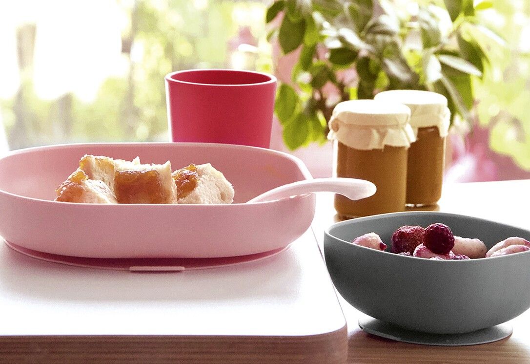 Piatto rosa con cibo, ciotola grigia con cibo, tazza rosa e cucchiaio. Su un tavolo.