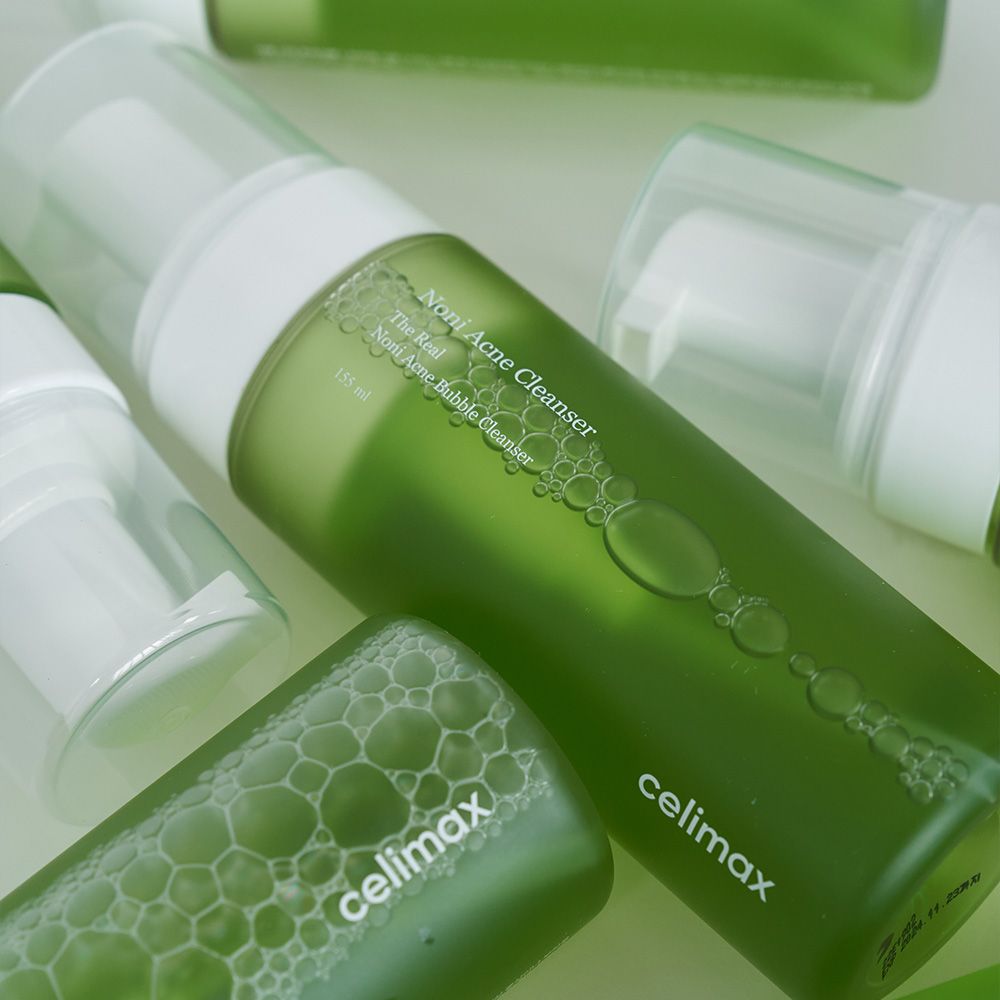 Molteplici flaconi di detergente schiumogeno anti-acne Celimax Noni, alcuni sdraiati, con contenuto verde ed erogatori a pompa.