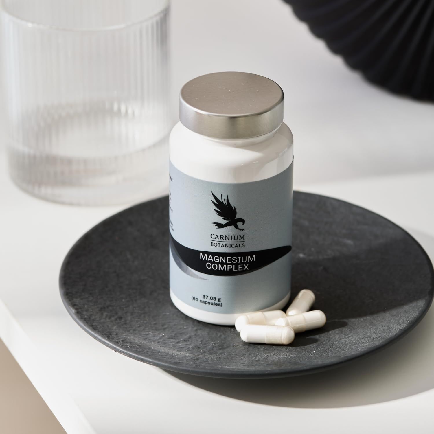 Flacone bianco con tappo argentato. Capsule su un piatto. Scritta: CARNIUM BOTANICALS, Magnesium Complex.