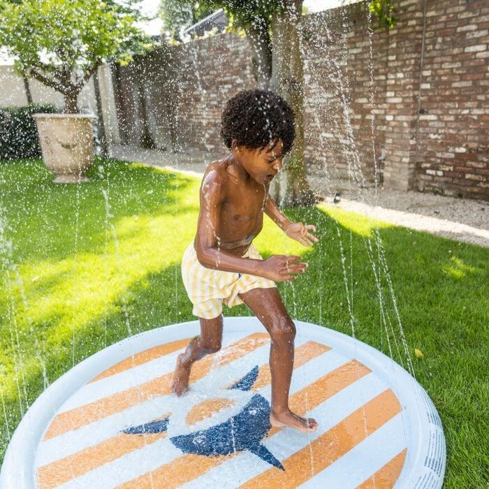 Bambino in pantaloncini a righe gialle corre su tappeto rotondo con motivo squalo. Spruzzi d'acqua.