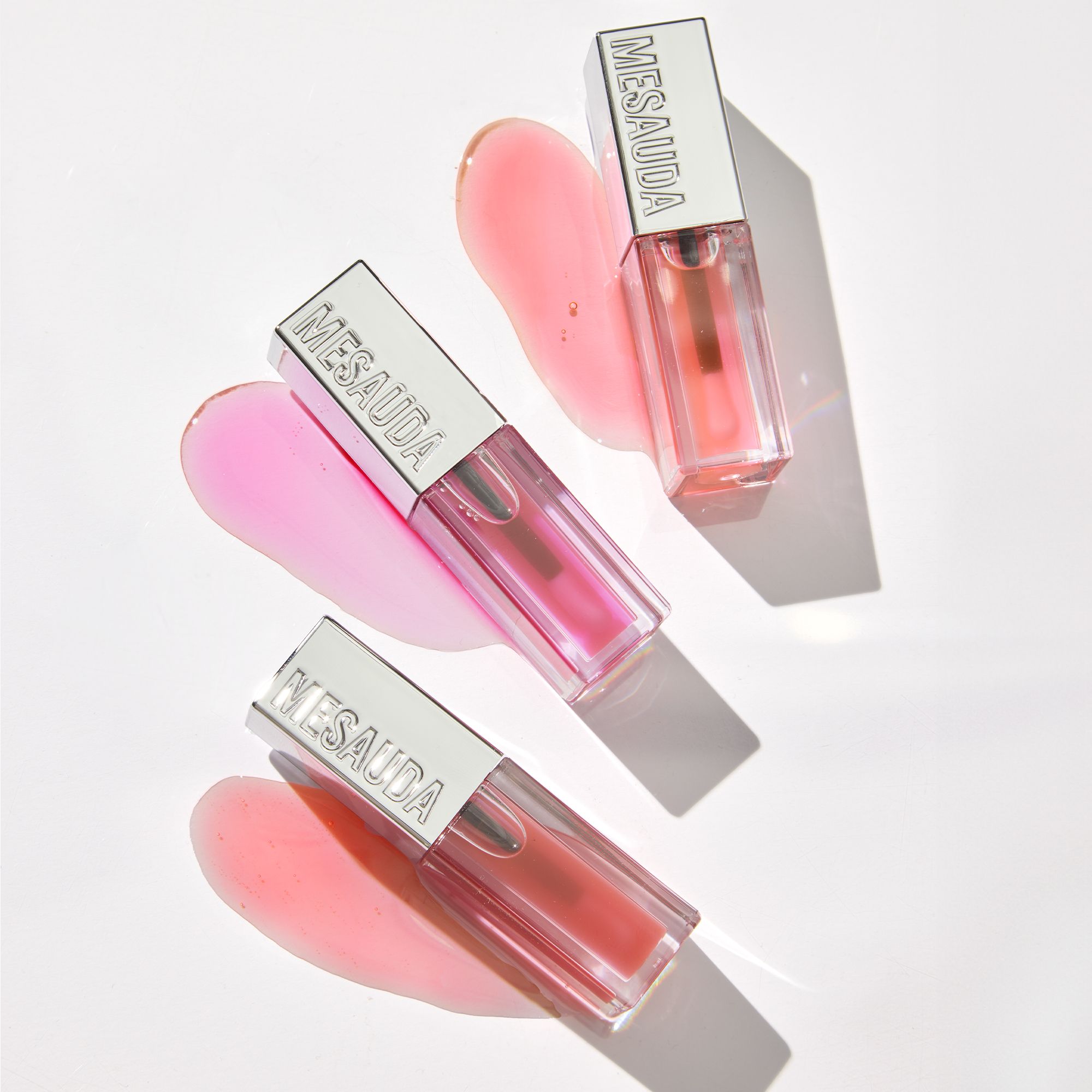 Tre flaconi di rossetto con tappi argentati recanti la scritta MESAUDA, disposti su tracce di prodotto rosa.