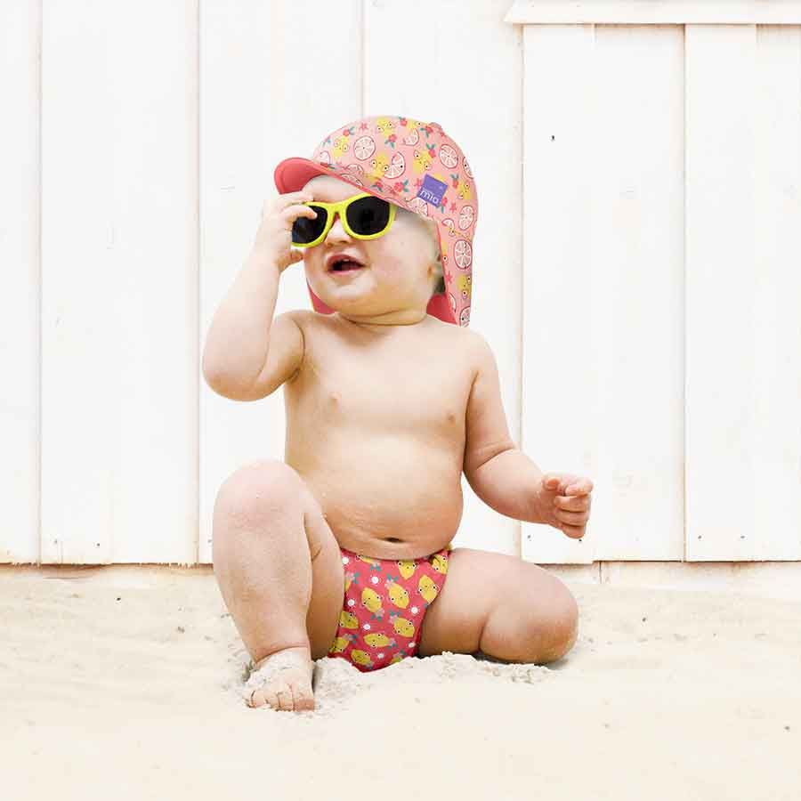 Bambino sulla spiaggia con cappello e occhiali da sole. Indossa costumino contenitivo con limoni. Seduto sulla sabbia.