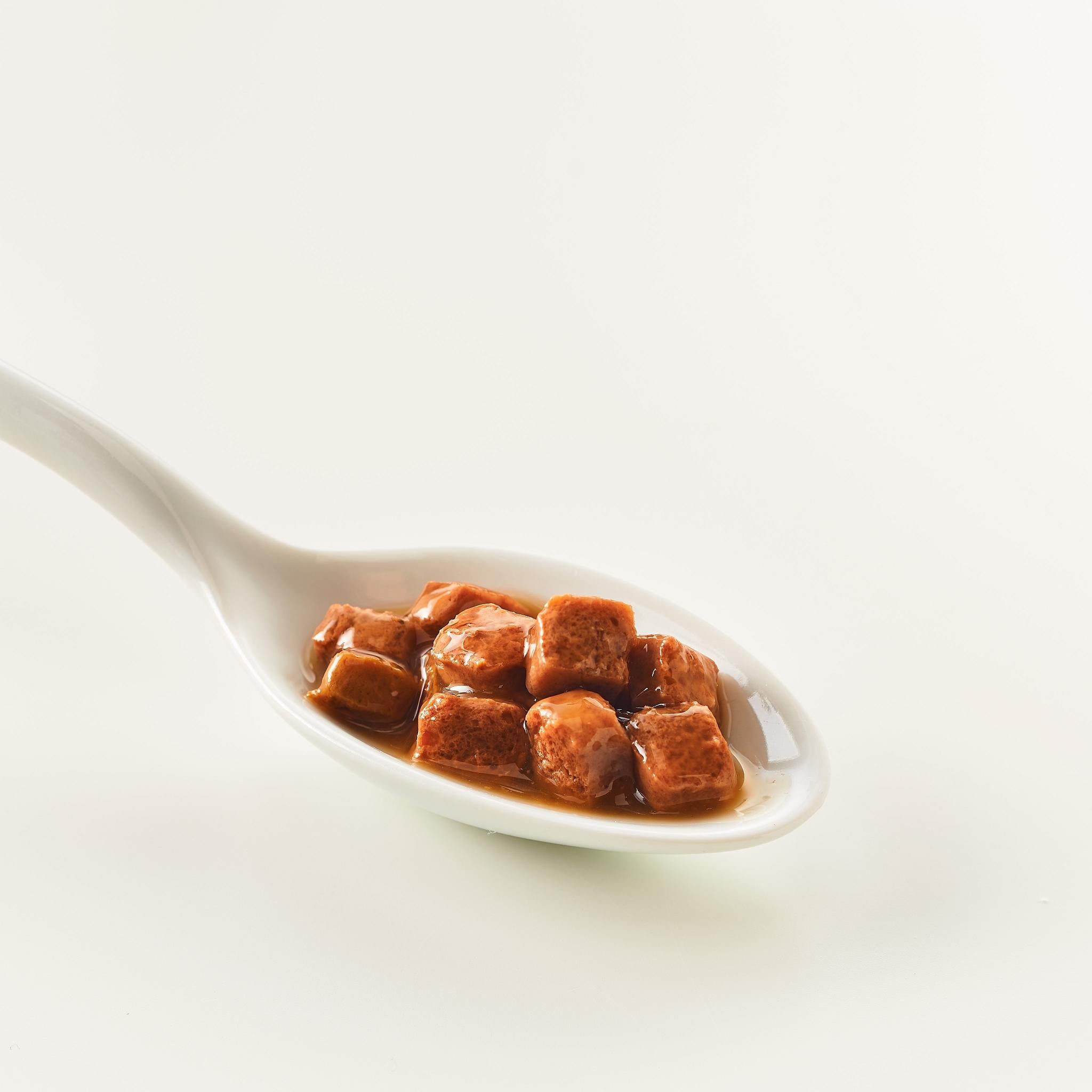 Cucchiaio con pezzi di cibo marroni e cubici in salsa. Primo piano su sfondo bianco.