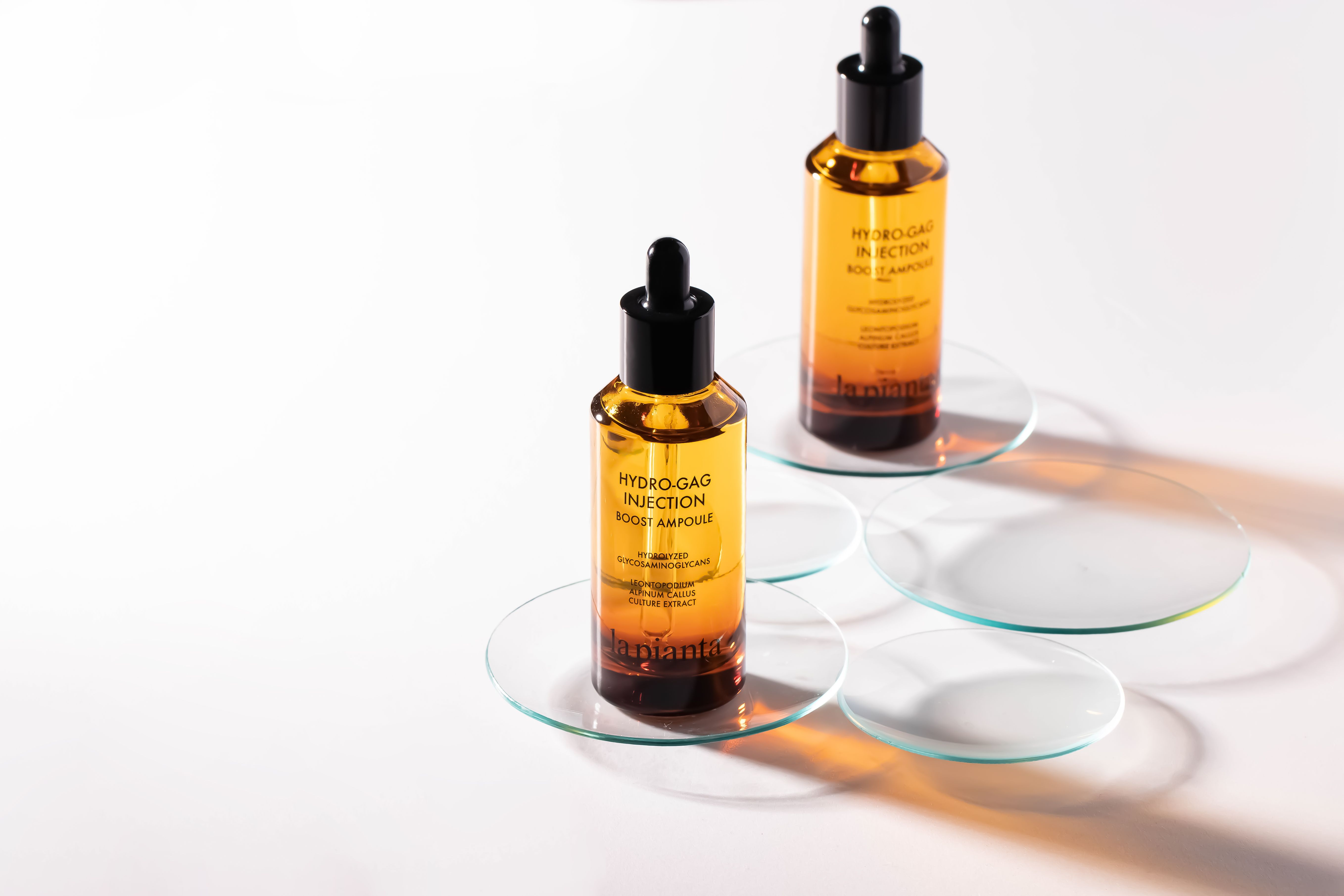 Due flaconi di vetro marrone con contagocce nero. Su sottobicchieri di vetro. Scritta: HYDRO-GAG INJECTION BOOST AMPOULE.