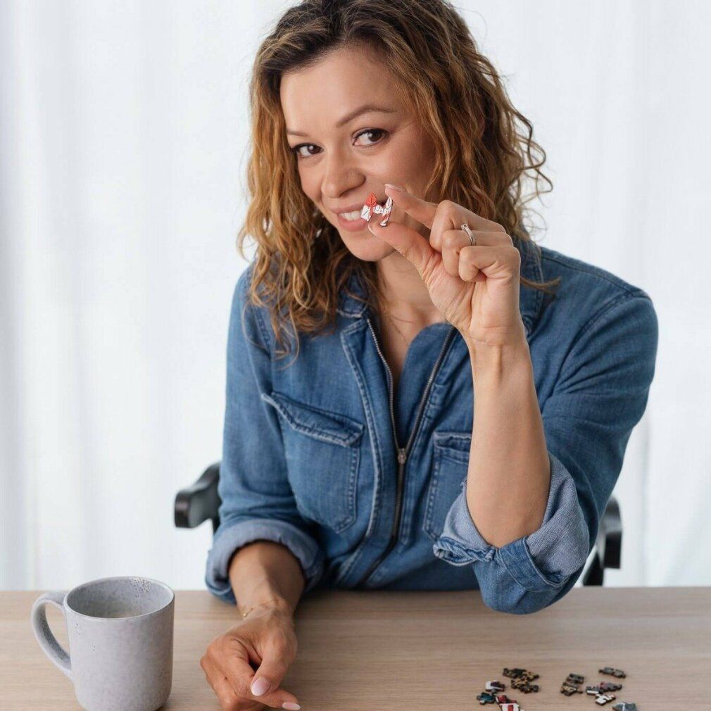 Donna tiene in mano piccoli pezzi di puzzle. Alcuni pezzi sono visibili. Una tazza è sul tavolo.