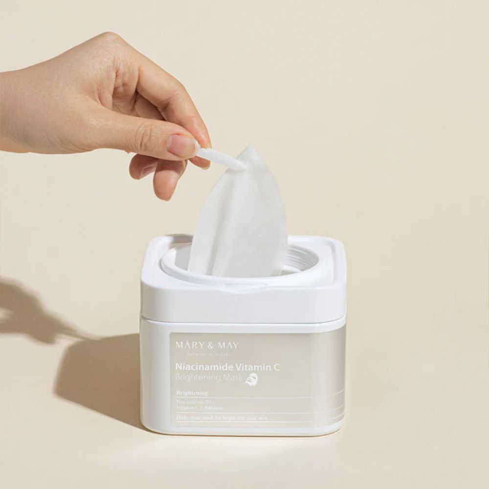 Mano che preleva una maschera da un contenitore bianco MARY & MAY Niacinamide Vitamin C Brightening Mask.