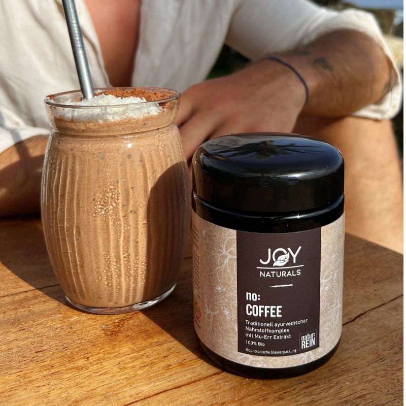 Bevanda in vetro con cannuccia e barattolo nero con 'JOY NATURALS no: COFFEE' su tavolo di legno. Uomo sullo sfondo.